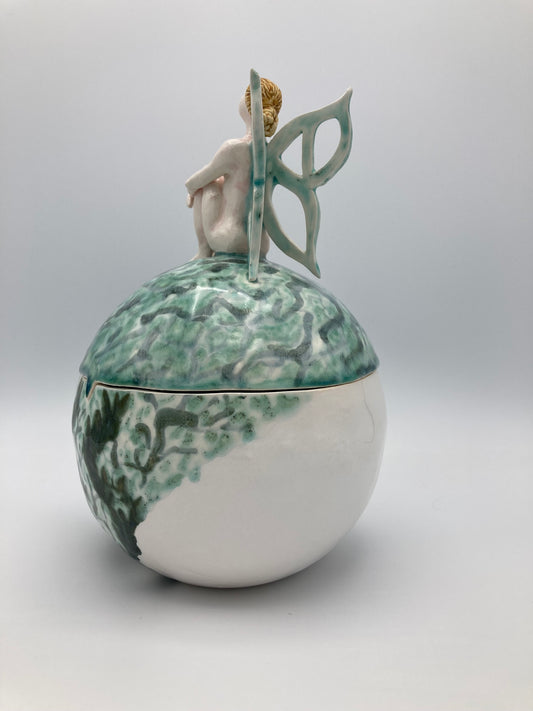 A picture of a sphere shape ceramic lidded box that would largely fill both your hands. A nude sculpture of a fairy sit on top of the box with her arms loosely wrapped around her knees in front of her. Her light green wings are gently opening behind her. The box is decorated in swirls of light transparent greens for the lid and darker greens and blues for the body of the box.