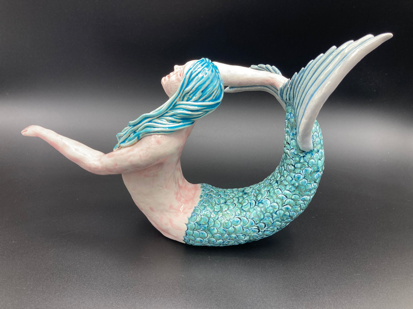 a photo of a gorgeous ceramic full body mermaid sculpture created by Shani and Anaick. Imagine that she was laid on her front and then lifted her torso up, reaching her left arm out in front of herself. Her beautiful blue-green scaled tail is curled up behind her and her right arm is stretched out backwards reaching for the top of her delicately fanned grey tail. Her long turquoise hair is draped delicately along the left side of her pretty face.