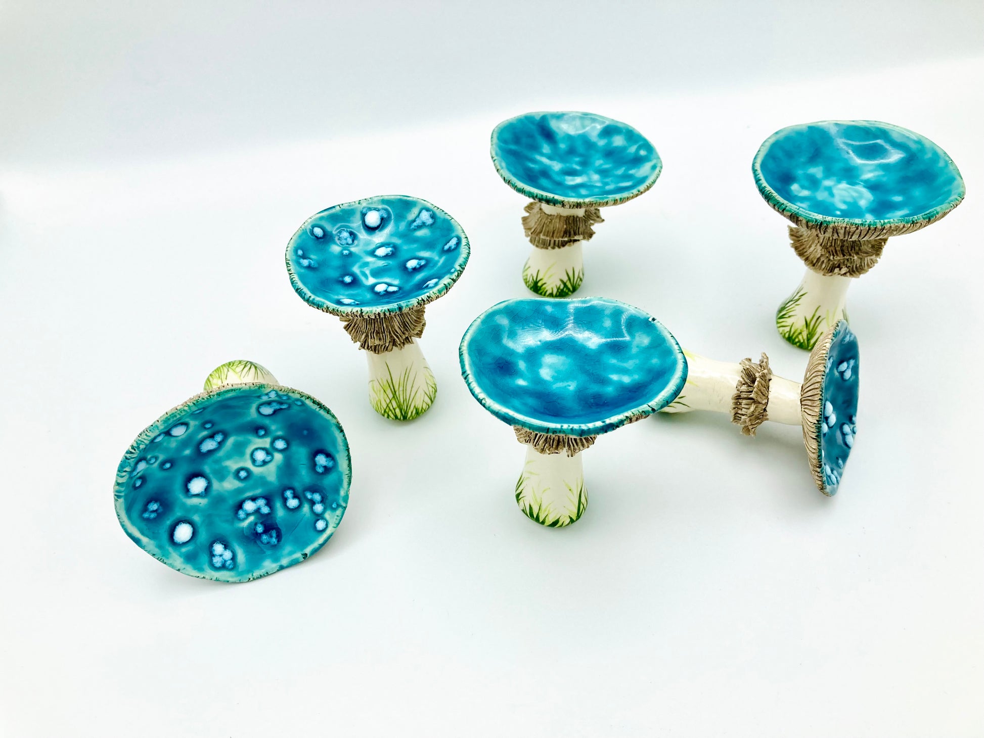 6 ceramic mushrooms with round, shallow pool like blue tops. they have light brown sculpted details on their stems which are also decorated with delicately painted grass like designs towards the bottom of the stems.