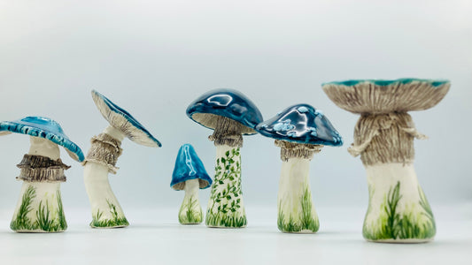 A series of 6 beautiful ceramics mushrooms with striking glossy blue tops. Their white stems are decorated with delicate green painted grass and leafy vines details. Each mushroom is around 10 cm high. They all have different shapes tops and one also has white dots like a toadstool mushroom. 
