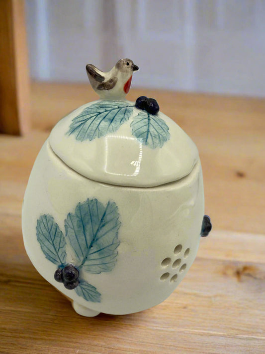A small round lidded pot that would fit in your hands put together and would hole a large full garlic clove. It is mainly light cream in colour with the print of blue bramble leaves decorating the sides and the lid.There are tiny 3D blackberries in each set of 3 leaves. Sat on the top of the lid is a tiny chirpy robin who is about to nibble at the 3 blackberries in front of him. The pot has little hole cut in its side in the shape of a flower to allow air through to keep garlic fresher for longer.