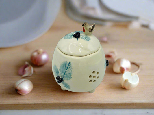 A small round lidded pot that would fit in your hands put together and would hole a large full garlic clove. It is mainly light cream in colour with the print of blue bramble leaves decorating the sides and the lid.There are tiny 3D blackberries in each set of 3 leaves. Sat on the top of the lid is a tiny chirpy robin who is about to nibble at the 3 blackberries in front of him. The pot has little hole cut in its side in the shape of a flower to allow air through to keep garlic fresher for longer.