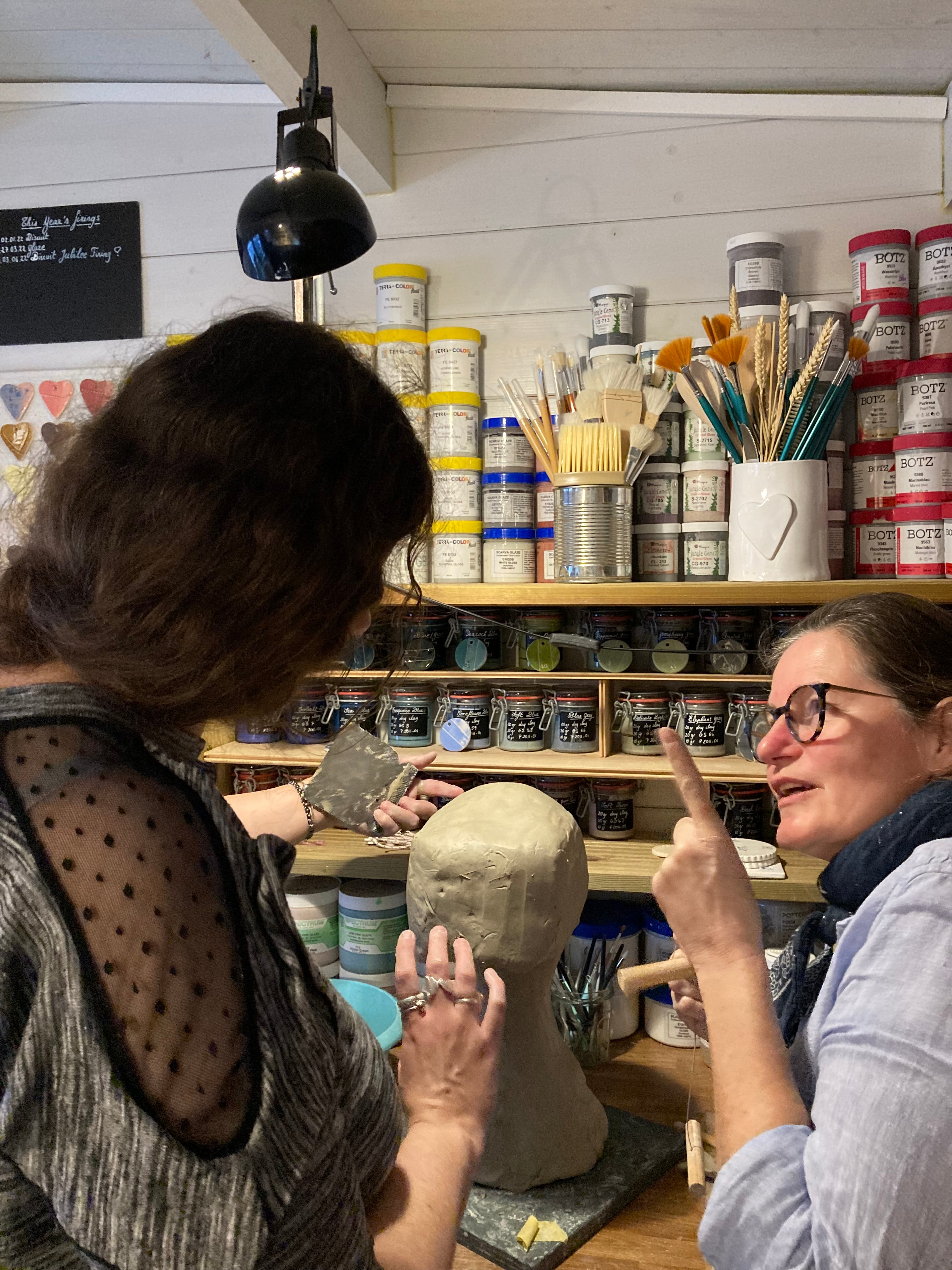 Shani and Anaick are working in the Happy studio. on the worktop, between them, sits a very rough sculpture of a ball of clay sitting on top of a neck shape piece of clay. one can guess that it will eventually become a portrait sculpture. The girls are deep in discussion, planning the next stages of the sculpting process. Shani is measuring the depth of the ball of clay between her hands and Anaick seems to have remembered something important and is waggling her finger in the air. lol