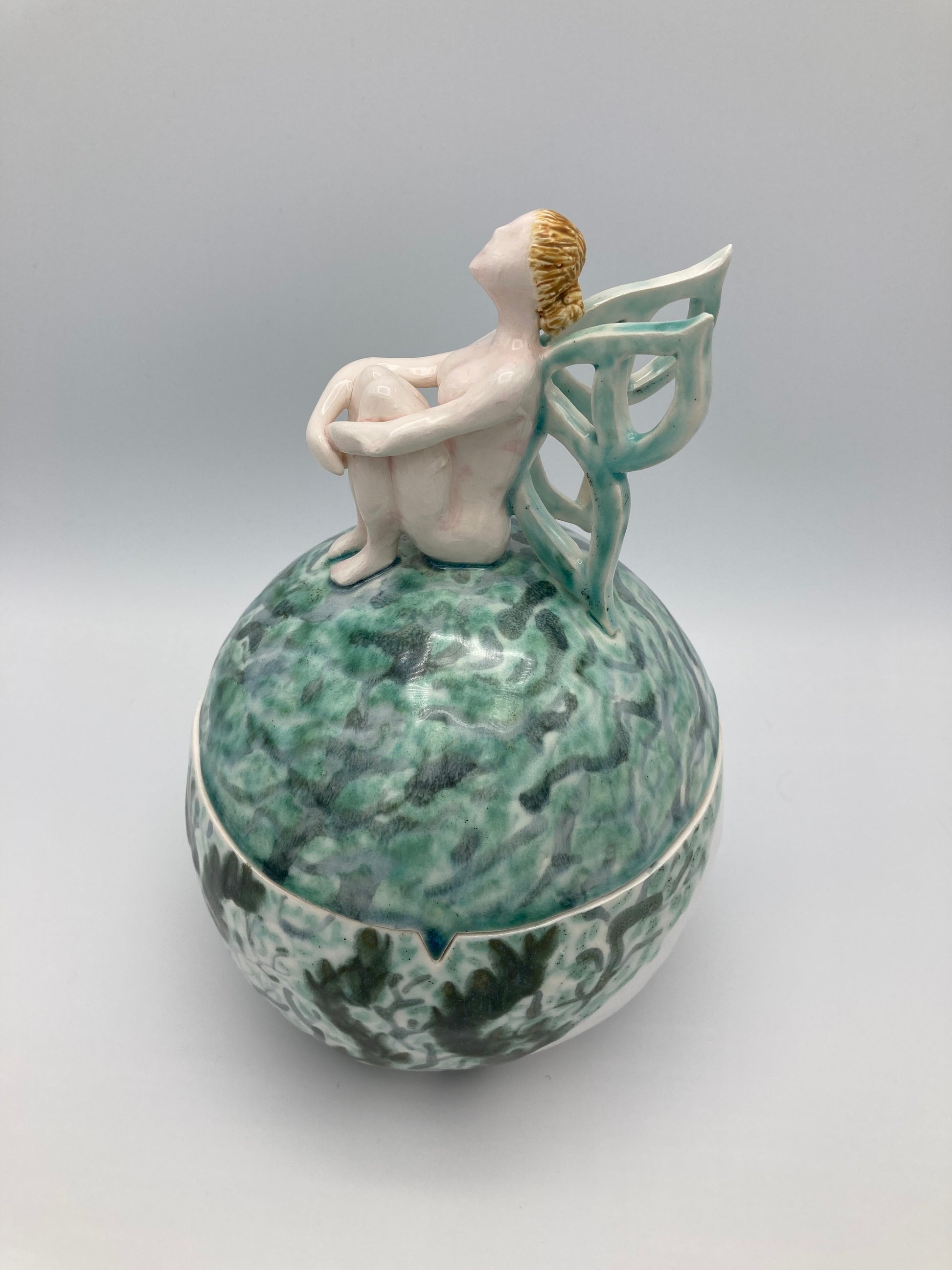 A picture of a sphere shape ceramic lidded box that would largely fill both your hands. A nude sculpture of a fairy sit on top of the box with her arms loosely wrapped around her knees in front of her. Her light green wings are gently opening behind her. The box is decorated in swirls of light transparent greens for the lid and darker greens and blues for the body of the box.