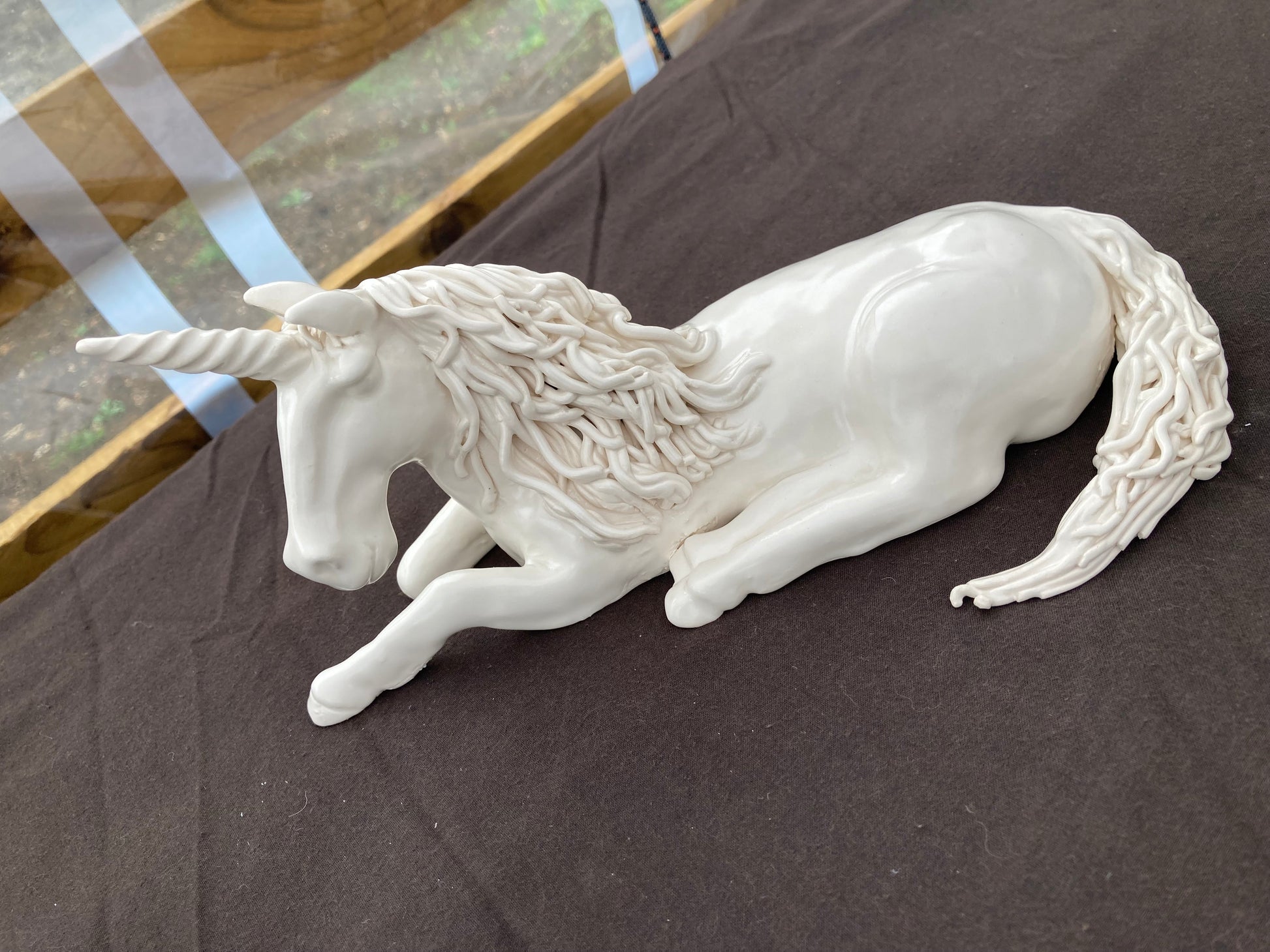 A white sculpture of a strong stallion laid down with his head up and one of his front leg folded in a manner suggesting that he is about to stand up. His mane and tail are made of hundreds of thinly rolled strips of clay and flow beautifully along his strong neck and on the ground around his hips.