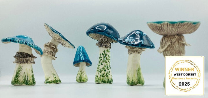 A series of 6 beautiful ceramics mushrooms with striking glossy blue tops. Their white stems are decorated with delicate green painted grass and leafy vines details. Each mushroom is around 10 cm high. They all have different shapes tops and one also has white dots like a toadstool mushroom. A sticker in the bottom right corner says: 'Winner West Dorset Awards 2025'