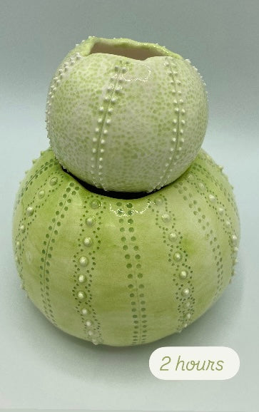 two decorative ceramic pots sculpted in the shape of urchins and glazed in different shades of light green are stacked on top of each other